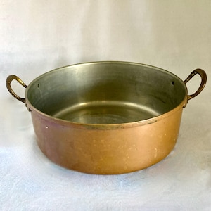 May include: A copper-colored cooking pot with two brass handles. The pot has a wide, shallow design with a silver-colored interior. The handles are curved and attached to the sides of the pot. The pot is likely used for cooking or serving food.