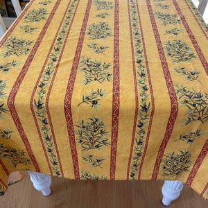 May include: Yellow tablecloth with vertical red stripes and olive branch patterns. The olive branches have green leaves and black olives. The tablecloth is on a white table.