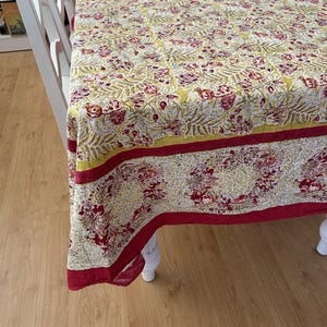 May include: A rectangular tablecloth featuring a repeating botanical pattern in shades of mustard yellow, deep red, and white. The fabric displays intricate floral and fern motifs with a solid red border along the edges, draped over a table.