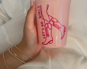 May include: Pink plastic cup with a pink graphic of a pair of cowboy boots and the text "Let's go girls".