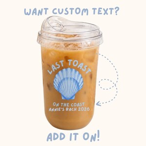May include: A clear plastic iced coffee cup with a lid. The cup has a light brown beverage inside with ice cubes. The cup has the text "LAST TOAST" and "ON THE COAST ANNIE'S BACH 2026" with a blue seashell graphic. The text "WANT CUSTOM TEXT?" and "ADD IT ON!" are also visible.