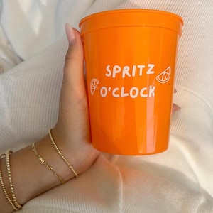 Puede incluir: Vaso de plástico naranja brillante con las palabras "SPRITZ O'CLOCK" en blanco y un gráfico de rodaja de cítrico. El vaso es sostenido por una persona que lleva pulseras doradas.
