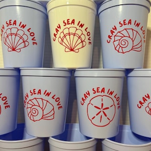 May include: A set of eight blue and white plastic cups with the phrase "Cray Sea in Love" and a red seashell design.