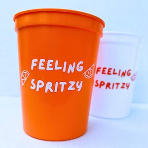 May include: Two plastic cups with the text "Feeling Spritzy" and a graphic of a lemon slice. The cup on the left is orange and the cup on the right is white.