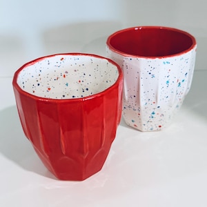 May include: Two ceramic cups with a faceted, geometric exterior. One cup is solid red on the outside with a white interior featuring blue and orange speckles. The second cup is white with blue and orange speckles on the outside and a solid red interior.