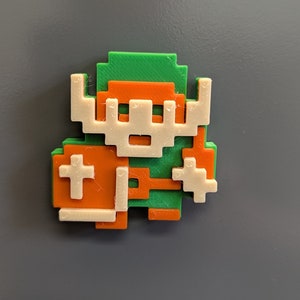 Puede incluir: Una figura impresa en 3D de un personaje de videojuego, hecha en verde, naranja y blanco. El personaje lleva una t&uacute;nica verde, pantalones naranjas y un casco blanco. El personaje sostiene una espada en una mano y un escudo en la otra.