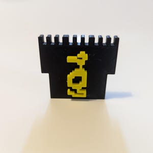 May include: A black, 3D-printed castle-shaped object with a yellow pixelated design on the front. The object has a rectangular base and a crenelated top, and the yellow design appears to be a stylized figure.