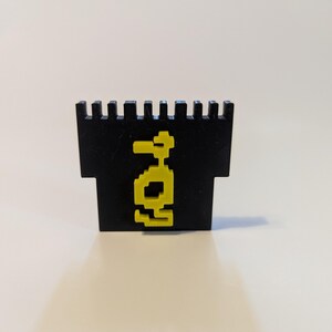 May include: A black, pixelated duck-shaped object with a yellow outline. The object has a rectangular base and a crenelated top. The duck graphic is centered on the front of the object.