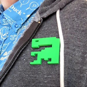 May include: A bright green, pixelated dinosaur pin is attached to a gray hooded sweatshirt. The pin is a solid, three-dimensional shape with a simple, blocky design. A blue button-down shirt with white and blue text is visible in the background.