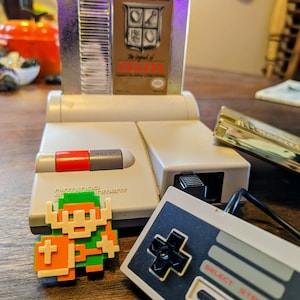 Puede incluir: Una consola Nintendo Entertainment System (NES) vintage de color gris y blanco. La consola tiene un cartucho de juego insertado, titulado "The Legend of Zelda". Un mando NES gris y negro se encuentra a la derecha de la consola. Una figura pixelada verde, naranja y blanca de Link del juego est&aacute; en primer plano.