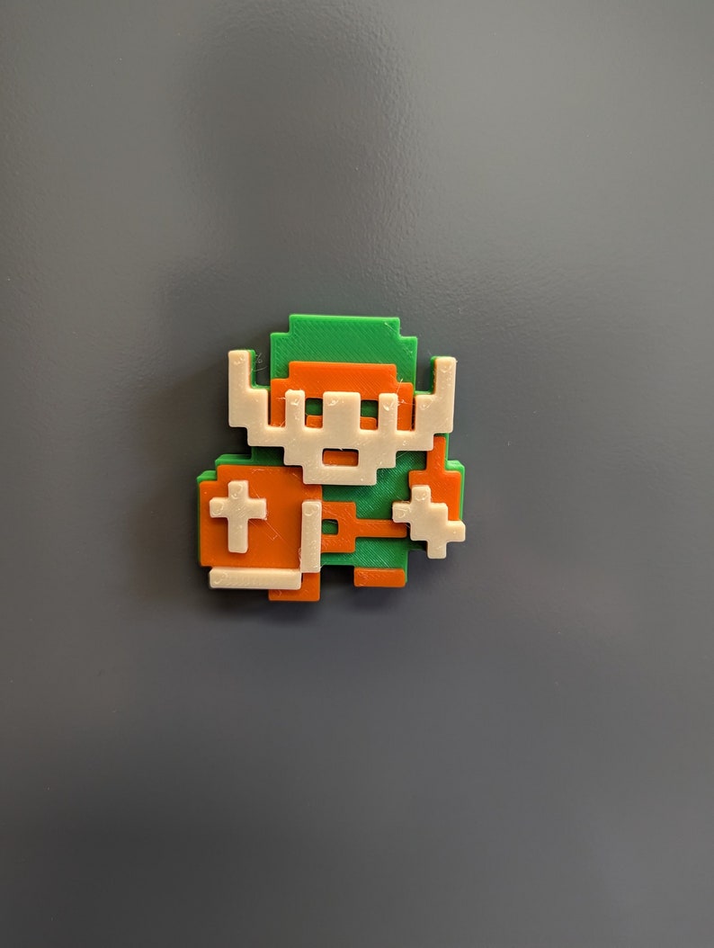 Puede incluir: Una figura impresa en 3D de un personaje de videojuego, hecha en verde, naranja y blanco. El personaje lleva una t&uacute;nica verde, pantalones naranjas y un casco blanco. El personaje sostiene una espada en una mano y un escudo en la otra.