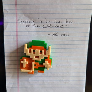 Puede incluir: Una figura impresa en 3D de color verde y naranja que representa a un personaje de videojuego. La figura est&aacute; sobre un trozo de papel rayado con el texto "Secret is in the tree at the dead-end" y "old man" escrito debajo.