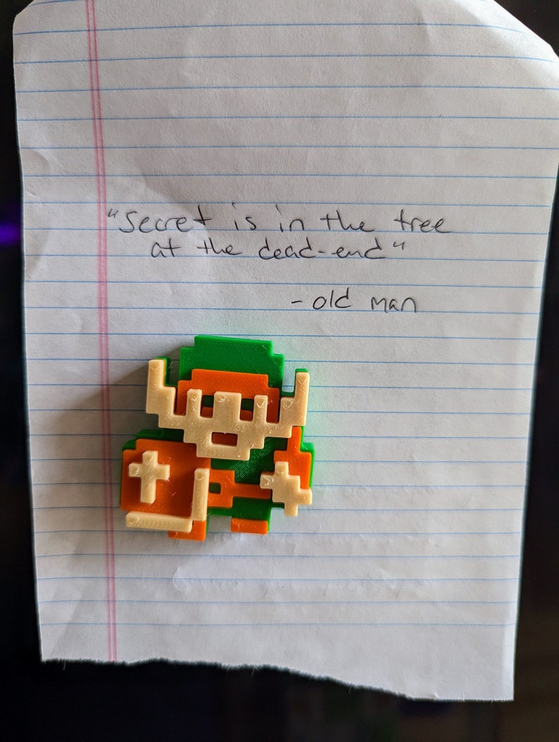 Puede incluir: Una figura impresa en 3D de color verde y naranja que representa a un personaje de videojuego. La figura est&aacute; sobre un trozo de papel rayado con el texto "Secret is in the tree at the dead-end" y "old man" escrito debajo.