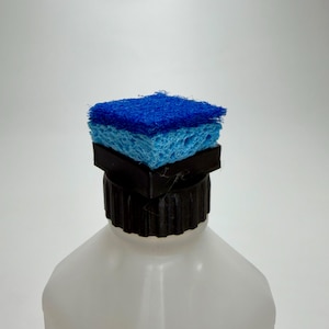 May include: A white plastic bottle with a black cap, topped with a blue and light blue sponge. The sponge is square-shaped and appears to be for cleaning purposes.