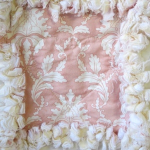 May include: A pink and white fabric square with a white ruffled border. The fabric has a damask pattern with white leaves on a pink background.