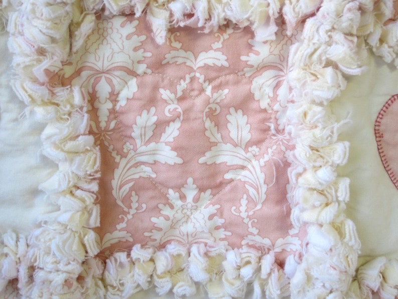 May include: A pink and white fabric square with a white ruffled border. The fabric has a damask pattern with white leaves on a pink background.