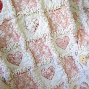 May include: A pink and white patchwork quilt with a heart design. The quilt is made of fabric with a floral pattern and has a white fringe around the edges.