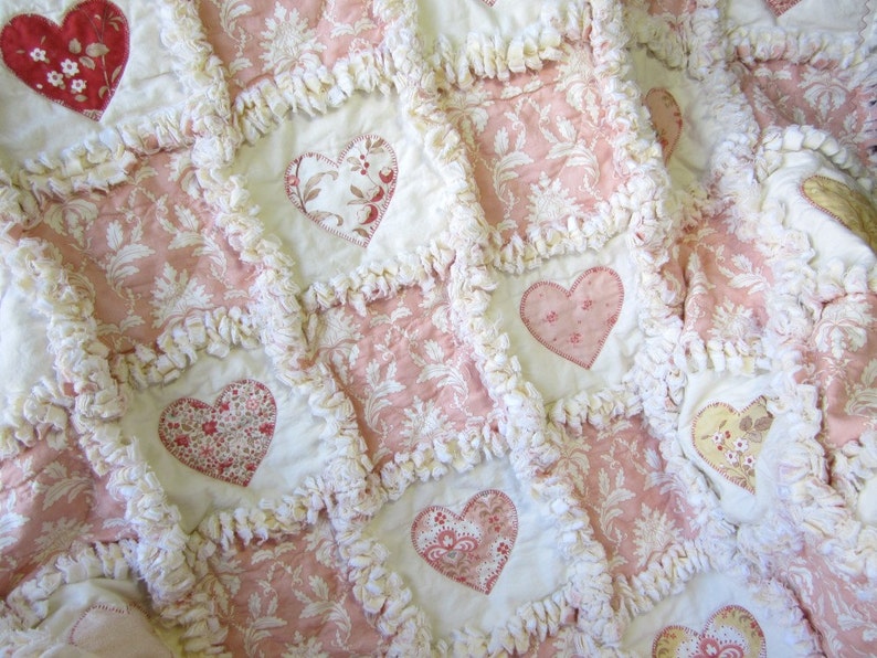 May include: A pink and white patchwork quilt with a heart design. The quilt is made of fabric with a floral pattern and has a white fringe around the edges.