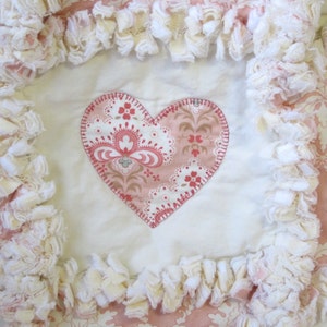 May include: A white and pink heart-shaped fabric patch with a floral pattern is sewn onto a white fabric background. The fabric background is surrounded by a white ruffled trim.