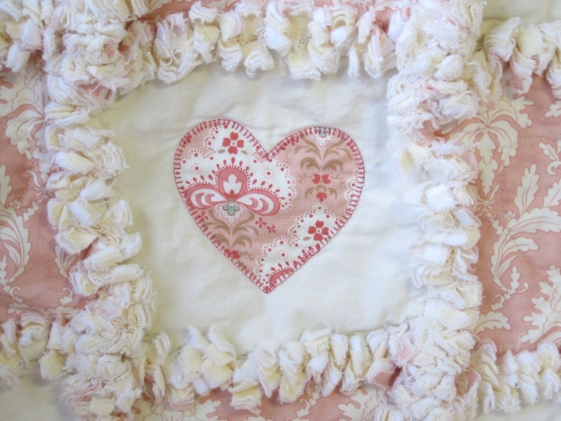 May include: A white and pink heart-shaped fabric patch with a floral pattern is sewn onto a white fabric background. The fabric background is surrounded by a white ruffled trim.