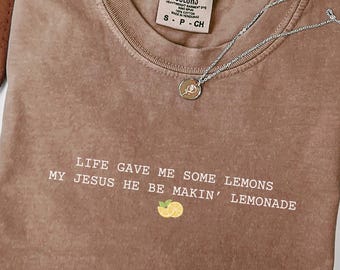 May include: Espresso-colored t-shirt with the text "LIFE GAVE ME SOME LEMONS MY JESUS HE BE MAKIN' LEMONADE" in white. A small lemon graphic is below the text. A silver necklace with a pendant is on the shirt.