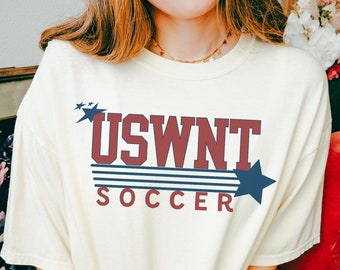 May include: Ivory t-shirt with a red and blue graphic that says "USWNT SOCCER" with stars and stripes.