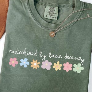 May include: Moss green t-shirt with the phrase "radicalized by basic decency" and colorful flower graphics. A gold necklace with a pendant rests on the shirt. A brown hat is visible in the background.