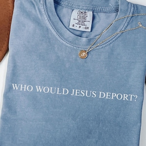 May include: A blue jean colored t-shirt with the text "WHO WOULD JESUS DEPORT?" in white. The shirt has a crew neck and short sleeves. A gold necklace with a pendant is draped on the shirt.