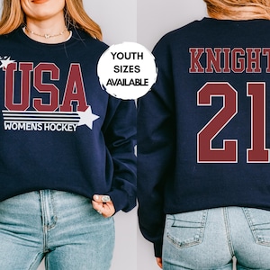 May include: Navy blue sweatshirt with "USA WOMENS HOCKEY" in maroon and white. The front design includes stars. The back features "KNIGHT 21" in maroon. Youth sizes available.