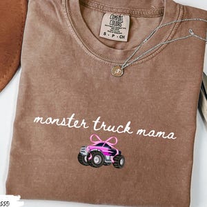 Puede incluir: Camiseta color espresso con las palabras "monster truck mama" en escritura blanca y un gr&aacute;fico de monster truck rosa y negro. Un collar plateado con un colgante est&aacute; sobre la camiseta.
