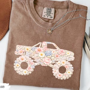 Op de afbeelding: Espresso-kleurig t-shirt met een bloemen monster truck ontwerp. De truck is versierd met roze, witte en blauwe bloemen. Het shirt heeft een ronde hals en korte mouwen. Een zilveren ketting met een kleine hanger ligt op het shirt.