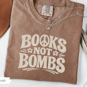 May include: Espresso-colored t-shirt with the text "BOOKS NOT BOMBS" in a retro font. The shirt features a peace sign and flower graphic. A silver necklace with a pendant is draped on the shirt.