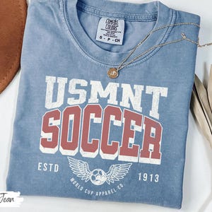 Może przedstawiać: Niebieska koszulka dżinsowa z napisem "USMNT SOCCER" w białych i czerwonych literach. Koszulka posiada piłkę nożną ze skrzydłami oraz napisy "ESTD 1913" i "WORLD CUP APPAREL CO." Złoty naszyjnik jest na koszulce.