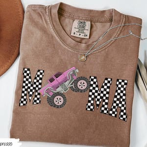 May include: Espresso-colored t-shirt with the word "MAMA" in a checkered pattern, featuring a pink monster truck graphic. The shirt has a crew neck and short sleeves. A silver necklace with a pendant is draped on the shirt.