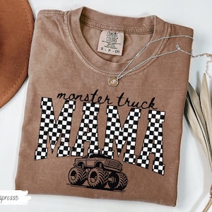 Op de afbeelding: Espresso-kleurig T-shirt met de tekst "monster truck MAMA" in zwart schrift en geblokte letters. Een monster truck-afbeelding staat onder de tekst. Een zilveren ketting met een hanger ligt op het shirt.