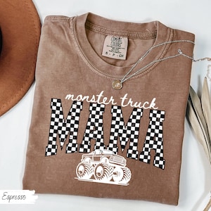 May include: Espresso-colored t-shirt with the words "monster truck MAMA" in white and a checkered pattern. A white monster truck graphic is below the text. The shirt has a crew neck and short sleeves. A silver necklace with a pendant is on the shirt.
