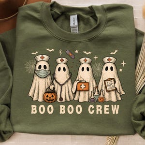 May include: Military green sweatshirt with a Halloween-themed design. The graphic features four ghost characters dressed as nurses, with the text "BOO BOO CREW" below. The ghosts are wearing medical accessories and holding a pumpkin.