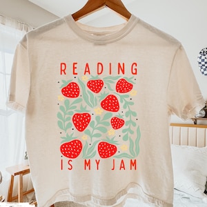 May include: Ivory-colored t-shirt with the text "READING IS MY JAM" in red. The shirt features a design of red strawberries, green leaves, and yellow flowers. The shirt is hanging on a wooden hanger.