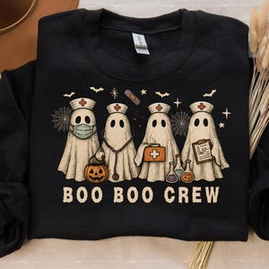 May include: Black sweatshirt featuring a Halloween-themed design. The graphic shows four ghost characters dressed as nurses, each with medical accessories. The text "BOO BOO CREW" is printed below the ghosts. A pumpkin is also included in the design.