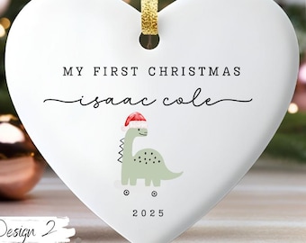 Pode incluir: Um enfeite de Natal branco em forma de coração com o texto "MY FIRST CHRISTMAS" e o nome "isaac cole". Apresenta um dinossauro verde usando um chapéu de Papai Noel e o ano de 2025. Uma fita dourada é anexada para pendurar.