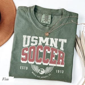 Może przedstawiać: Koszulka USMNT Soccer w kolorze mchu z białymi i bordowymi literami. Koszulka przedstawia piłkę nożną ze skrzydłami i napisem "World Cup Apparel Co. ESTD 1913". Złoty naszyjnik spoczywa na koszulce.