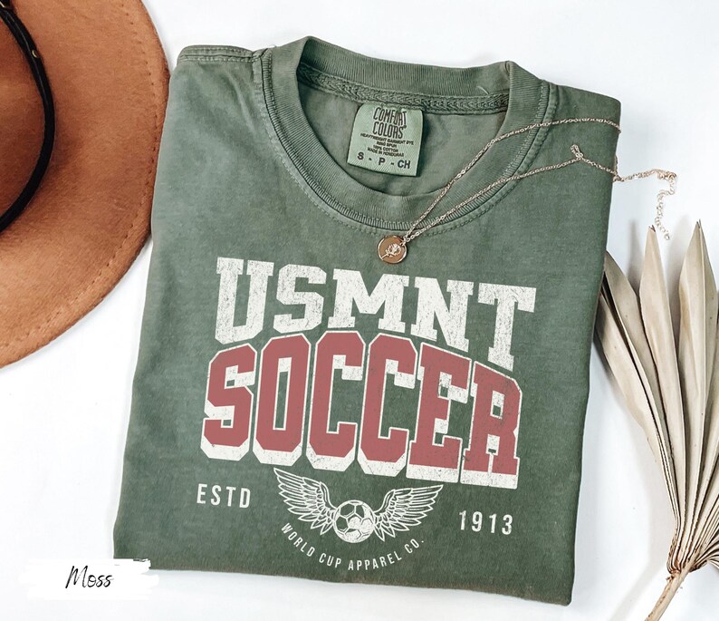 Może przedstawiać: Koszulka USMNT Soccer w kolorze mchu z białymi i bordowymi literami. Koszulka przedstawia piłkę nożną ze skrzydłami i napisem "World Cup Apparel Co. ESTD 1913". Złoty naszyjnik spoczywa na koszulce.