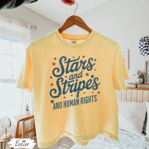 Puede incluir: Camiseta amarilla con el texto "Stars and Stripes AND HUMAN RIGHTS" en letras azul marino y estrellas rojas. La camiseta est&aacute; colgada en una percha de madera.