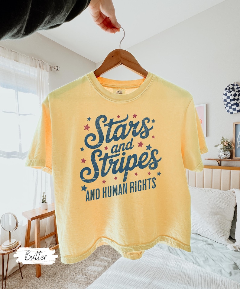 Puede incluir: Camiseta amarilla con el texto "Stars and Stripes AND HUMAN RIGHTS" en letras azul marino y estrellas rojas. La camiseta est&aacute; colgada en una percha de madera.