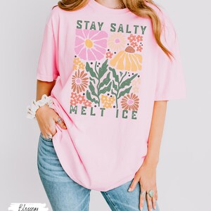 Op de afbeelding: Lichtroze t-shirt met de tekst "STAY SALTY MELT ICE" in groen en een bloemenontwerp in roze, geel en bruin. Het shirt is een casual stijl met korte mouwen.