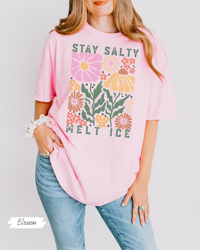Op de afbeelding: Lichtroze t-shirt met de tekst "STAY SALTY MELT ICE" in groen en een bloemenontwerp in roze, geel en bruin. Het shirt is een casual stijl met korte mouwen.