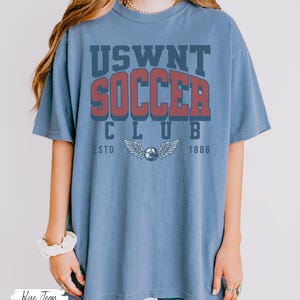 Puede incluir: Una camiseta azul con el texto "USWNT SOCCER CLUB ESTD 1886" impreso en la parte delantera. El texto est&aacute; en una fuente de estilo vintage y presenta un bal&oacute;n de f&uacute;tbol con alas.