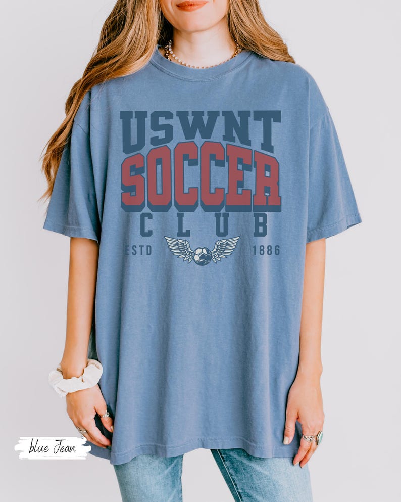 Puede incluir: Una camiseta azul con el texto "USWNT SOCCER CLUB ESTD 1886" impreso en la parte delantera. El texto est&aacute; en una fuente de estilo vintage y presenta un bal&oacute;n de f&uacute;tbol con alas.