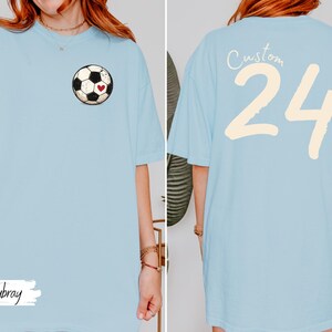Op de afbeelding: Lichtblauw T-shirt met een voetbalafbeelding en een rood hart op de voorkant. De achterkant van het shirt toont het nummer 24 in cr&egrave;me, met het woord "Custom" erboven. Het woord "Chanbray" staat op de voorkant.