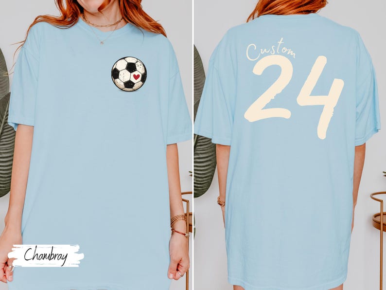 Op de afbeelding: Lichtblauw T-shirt met een voetbalafbeelding en een rood hart op de voorkant. De achterkant van het shirt toont het nummer 24 in cr&egrave;me, met het woord "Custom" erboven. Het woord "Chanbray" staat op de voorkant.